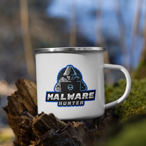 Könnte beinhalten: Eine weiße Emailletasse mit einem blauen und schwarzen Logo, auf dem "Malware Hunter" steht. Das Logo zeigt eine Person mit Kapuze, die vor einem Laptop mit einem Totenkopf auf dem Bildschirm sitzt.