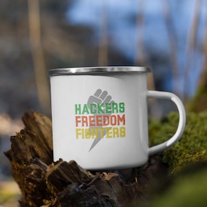 Könnte beinhalten: Weiße Emailtasse mit einem schwarzen Faust-Motiv und dem Text "Hackers Freedom Fighters" in Grün, Rot und Gelb.