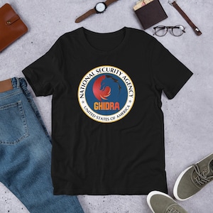 Könnte beinhalten: Schwarzes T-Shirt mit einem Grafikdruck eines roten, gelben und blauen Logos mit der Aufschrift "National Security Agency" und "GHIDRA" mit einem Stern auf jeder Seite des Textes. Das Logo ist von einem goldenen Kreis mit dem Text "United States of America" umgeben.
