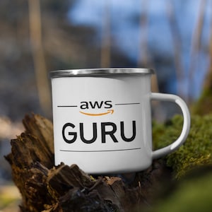 May include: White enamel mug with a black and orange logo that says "aws GURU" on it. The mug is sitting on a piece of wood with moss growing on it.