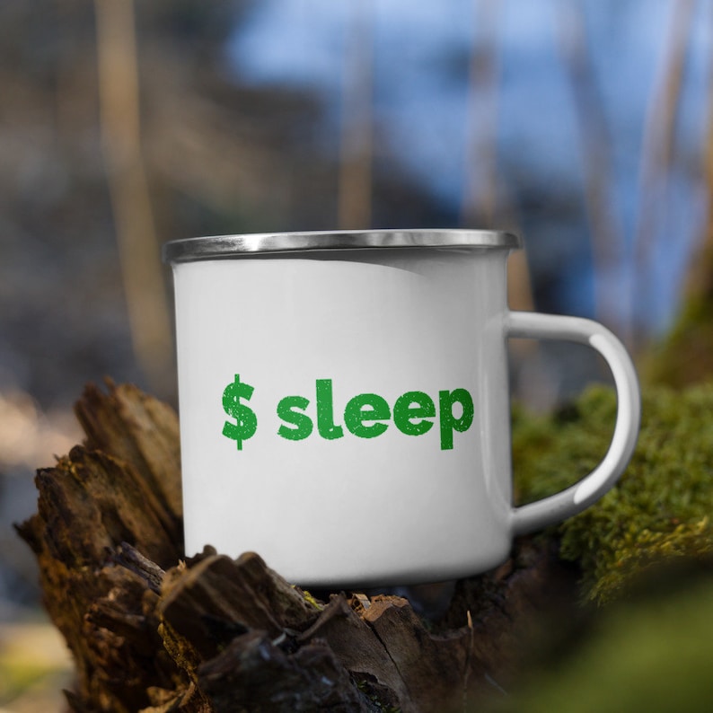 Peut inclure: Mug de camping en &eacute;mail blanc avec un dollar vert et le mot "sleep" imprim&eacute; dessus. Le mug est pos&eacute; sur un morceau de bois recouvert de mousse.