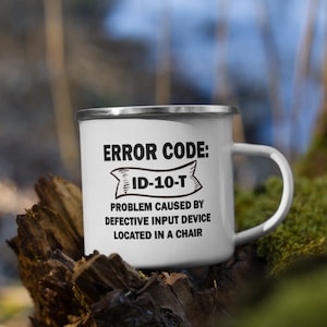 Könnte beinhalten: Eine weiße Emailletasse mit einer schwarz-weißen Grafik, auf der "ERROR CODE: ID-10-T PROBLEM CAUSED BY DEFECTIVE INPUT DEVICE LOCATED IN A CHAIR" steht.