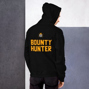 Könnte beinhalten: Schwarzer Hoodie mit einem Käfer-Motiv und dem Text "BOUNTY HUNTER" in Gelb.