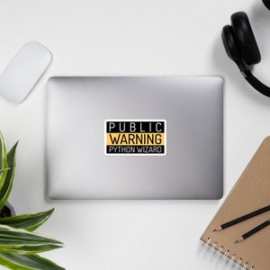 K&ouml;nnte beinhalten: Ein schwarzes und gelbes Warnschild mit dem Text "PUBLIC WARNING PYTHON WIZARD" auf einem Laptop.