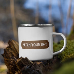 Könnte beinhalten: Weiße Emaille-Campingtasse mit einem braunen Patch, auf dem "PATCH YOUR SH*T" steht. Die Tasse steht auf einem Stück Holz mit Moos.