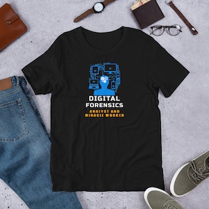 Könnte beinhalten: Schwarzes T-Shirt mit einer blauen Grafik eines Kopfes, der mit digitalen Geräten gefüllt ist, und dem Text "DIGITAL FORENSICS ANALYST AND MIRACLE WORKER" in Weiß und Gelb. Das T-Shirt liegt auf einer grauen Oberfläche.