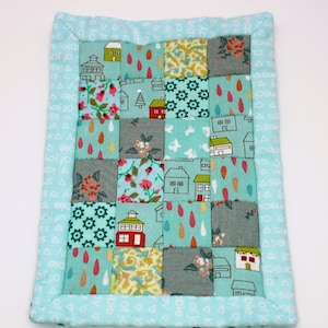 Könnte beinhalten: Ein kleines, türkisfarbenes, gestepptes Quadrat mit einem Patchwork-Muster aus Häusern, Bäumen und Regentropfen. Der Rand ist hellblau mit einem weißen Muster.