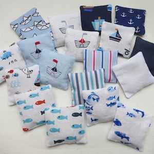 Op de afbeelding: Een verzameling kleine, vierkante stoffen zitzakken met nautische patronen. Ontwerpen omvatten zeilboten, ankers, vissen en geometrische strepen in blauw, rood en wit. Sommige zakken zijn effen van kleur.