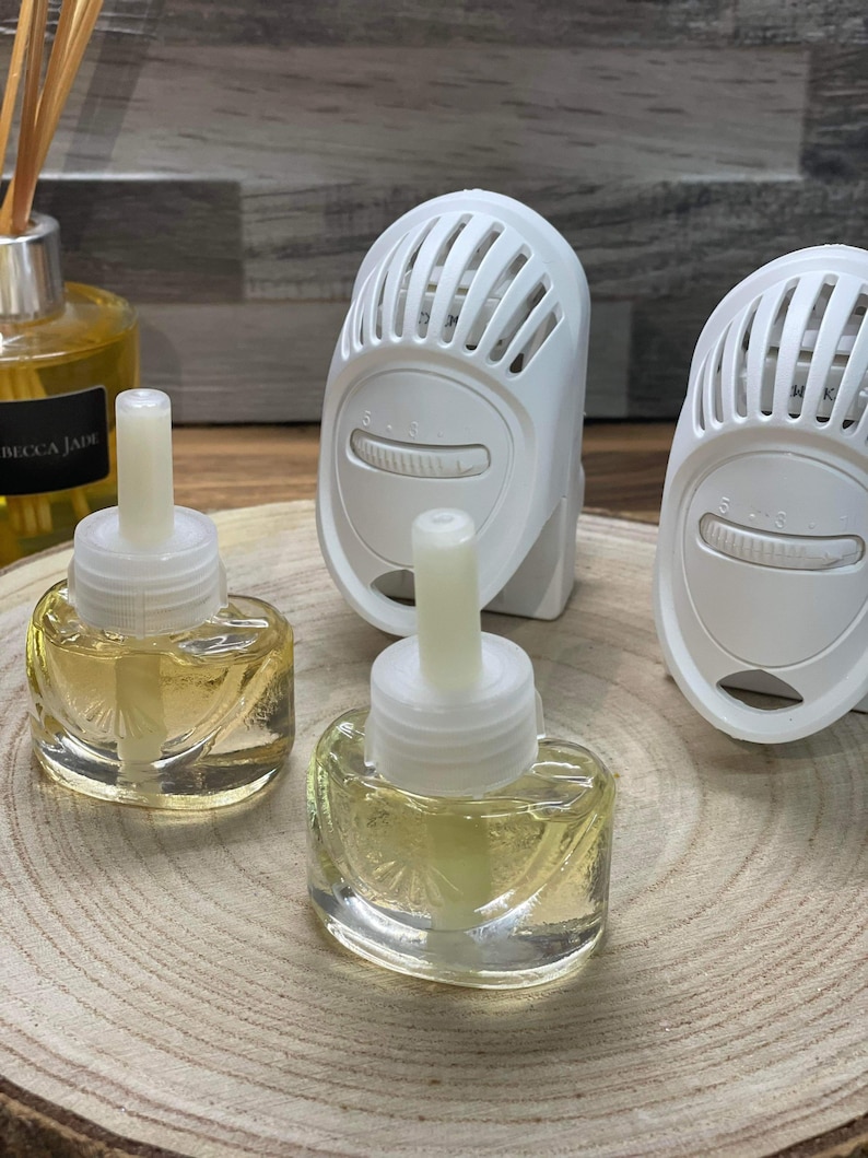 Op de afbeelding: Twee doorzichtige glazen flessen met gele vloeistof en witte doppen, die op een houten oppervlak staan. Op de achtergrond staan twee witte elektrische diffusers met een draaiknop om de instellingen aan te passen.