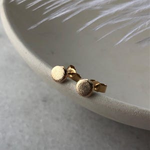 以下が含まれることがあります： 丸いテクスチャデザインのゴールドトーンのスタッドピアスのペア。ピアスは白い表面に表示されています。