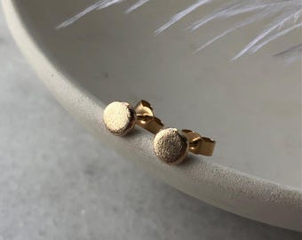 May include: A pair of gold-toned stud earrings with a round, textured design. The earrings are displayed on a white surface.