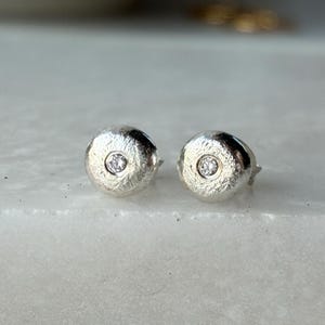 以下が含まれることがあります： テクスチャ加工されたつや消しの金属表面を持つ、円形のシルバー製スタッドピアス一対。それぞれの中心には、小さな透明なラウンドカットの宝石がセットされており、ドーム型の形状と金属的な光沢のある仕上げが特徴です。