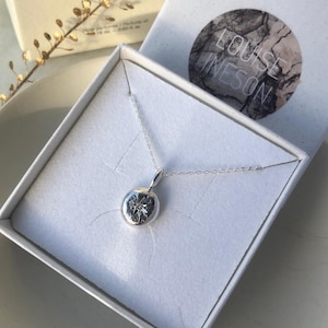 May include: A silver necklace with a round pendant featuring a star design. The necklace is in a white box with the name "LOUISE INESON" printed on the lid.