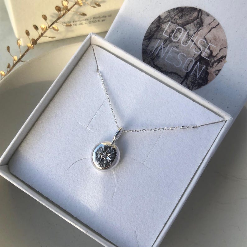 May include: A silver necklace with a round pendant featuring a star design. The necklace is in a white box with the name "LOUISE INESON" printed on the lid.