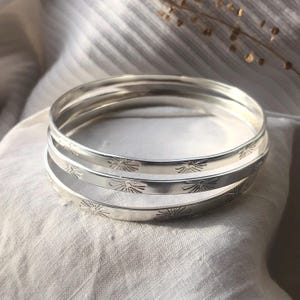 May include: A set of four silver bangles. Each bangle is engraved with a repeating floral pattern. The bangles are stacked on top of each other, resting on a white, textured surface. The bangles have a polished, reflective surface.