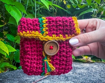 May include: A magenta crochet pouch with yellow and green detailing. It has a wooden button fastening and a tassel. The item is held by a person's hand against a backdrop of green foliage. The pouch is likely handmade.