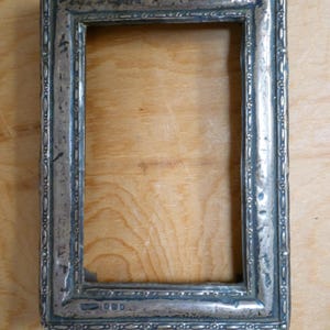 May include: An antique silver picture frame, rectangular in shape. The frame features a detailed border with a repeating pattern of small ovals and dots. The silvered surface shows signs of age and wear, with a slightly tarnished appearance.