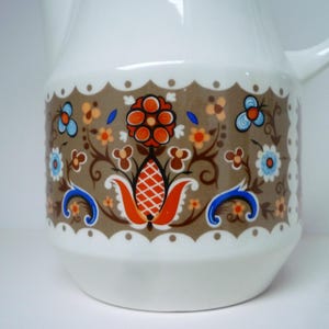 May include: White ceramic jug with a brown, blue and orange floral design. The jug has a scalloped edge and a handle.