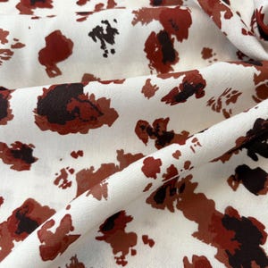 May include: White fabric featuring a repeating pattern of abstract shapes in shades of brown and dark brown. The fabric is folded, revealing its texture and drape. The pattern has a spotted appearance.