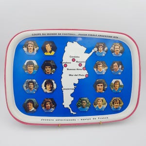 Op de afbeelding: Een rechthoekig metalen dienblad met een roze rand, een blauwe achtergrond, een witte kaart van Argentinië met rode stadsmarkeringen en zestien zeshoekige portretten van Franse voetbalspelers van het WK 1978, met Franse tekst.
