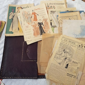 Puede incluir: Una colección de patrones de costura de papel vintage e ilustraciones de moda con bocetos de vestidos y lencería, junto a una carpeta de cuero marrón oscuro con un borde cosido y un cierre a presión, todo sobre una superficie blanca.