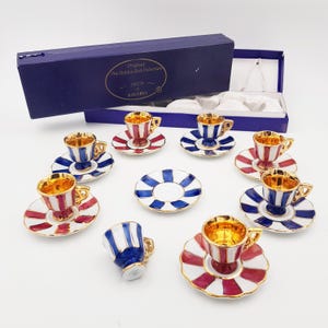 Op de afbeelding: Een set van acht miniatuur espresso kopjes en schoteltjes in een blauwe doos. De kopjes en schoteltjes zijn versierd met rode, witte en blauwe strepen en gouden accenten. De doos heeft de tekst "Original The Golden Red Collection" en "Bavaria" erop.