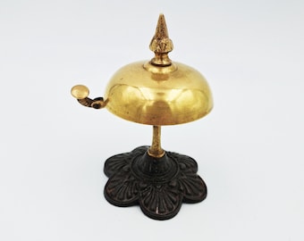 May include: A brass desk bell featuring a polished gold-tone dome with a pointed finial top and a small lever on the side. The bell is supported by a dark, textured, flower-shaped base that provides a stable foundation for the metallic structure.