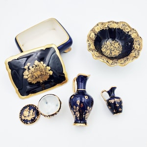 Op de afbeelding: Een verzameling donkerblauw porseleinen voorwerpen met gouden accenten, waaronder een rechthoekige doos met deksel, een kom met geschulpte rand, een klein potje met deksel en twee kannen van verschillende grootte, voorzien van gouden bloemmotieven.