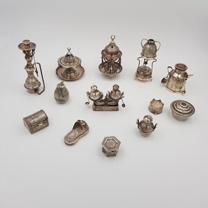 Puede incluir: Una colección de miniaturas de plata antiguas, que incluyen una cachimba, quemadores de incienso, un pequeño cofre, una zapatilla y varios recipientes decorativos. Los artículos están intrincadamente detallados y dispuestos sobre una superficie blanca.