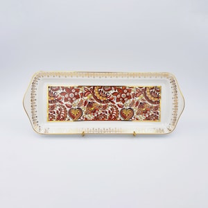 Peut inclure: Un plateau de service rectangulaire en céramique blanche avec un panneau central orné d'un motif dense et complexe de cachemire et de fleurs dans des tons rouille, orange et marron. Le bord présente une frise dorée et des finitions dorées.
