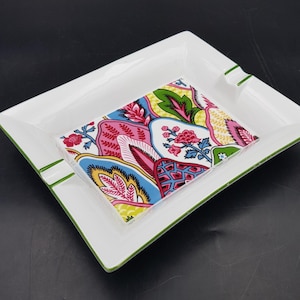 Könnte beinhalten: Weißer quadratischer Aschenbecher mit grünem Rand und einem bunten Fliesen-Einsatz mit Blumenmuster. Die Fliese zeigt rosa, blaue, gelbe und grüne Blumendesigns. Der Aschenbecher hat zwei grüne Rillen zum Ablegen von Zigaretten.