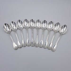 Pode incluir: Um conjunto de onze colheres de metal prateado dispostas num arco horizontal. Cada colher apresenta uma taça oval polida e um cabo fino que termina numa extremidade alargada decorada com um motivo de concha em relevo na base.