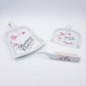 Peut inclure: Un ensemble en céramique blanche composé de deux pelles et d'une brosse, décorés de motifs de fleurs de cerisier roses. La grande pelle porte l'inscription "Ramasse Couverts" et la petite pelle porte l'inscription "Ramasse Miettes".