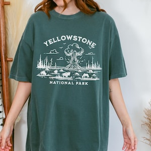 Puede incluir: Una camiseta de gran tamaño de color azul verdoso con un estampado gráfico blanco de un géiser en erupción en el Parque Nacional de Yellowstone. El texto "Yellowstone National Park" está impreso debajo del gráfico.