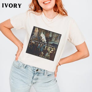 May include: A white t-shirt with a graphic print of a painting. The painting depicts a group of people in a historical setting. The text "Girls Will Be Girls" is printed on the shirt.