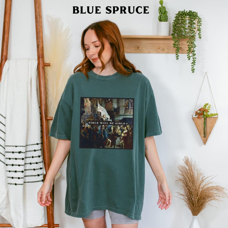May include: A teal blue t-shirt with a graphic print of a painting. The painting depicts a group of people in a room with the text "Girls Will Be Girls" in the center.