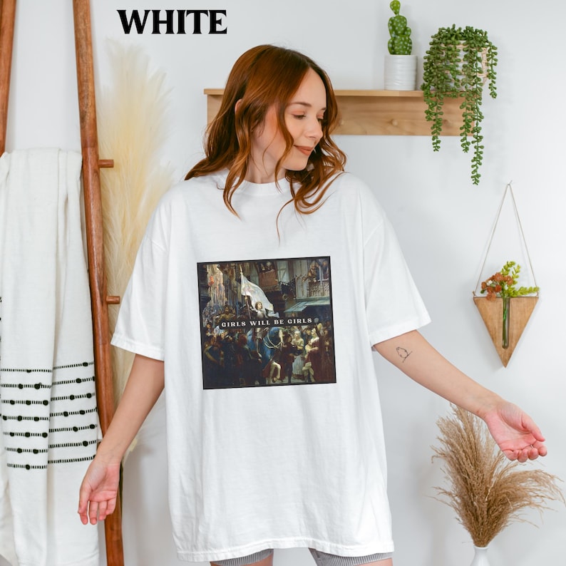 May include: White oversized t-shirt with a graphic print of a painting. The painting depicts a group of people in a street scene. The text "Girls Will Be Girls" is printed on the shirt.