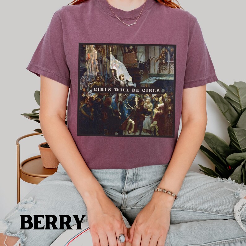 May include: A maroon t-shirt with a graphic print of a painting. The painting depicts a group of people in a historical setting. The text "Girls Will Be Girls" is printed across the bottom of the painting.