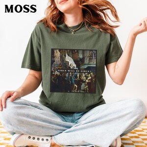 May include: A green t-shirt with a graphic print of a painting. The painting depicts a group of people in a historical setting. The text "Girls Will Be Girls" is printed on the shirt.