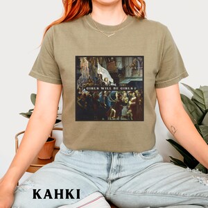 May include: A khaki t-shirt with a graphic print of a painting depicting a group of people. The text "Girls Will Be Girls" is printed on the graphic.