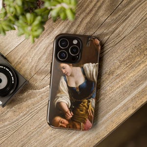 May include: A phone case featuring a painting of a woman holding a knife to a man's throat. The woman is wearing a blue and gold dress and the man is wearing a brown and pink shirt. The painting is in a dark, dramatic style.