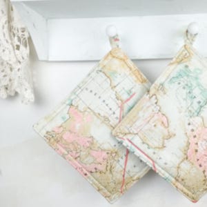 May include: Two square pot holders with a vintage map design. The pot holders feature a cream background with a map print in shades of pink, green, and tan. They are hanging on white hooks.