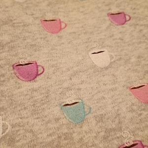 May include: Close-up of a gray fabric with an embroidered pattern of coffee cups. The cups are in various colors, including pink, blue, and white, with steam rising from them. The design is repeated across the fabric.