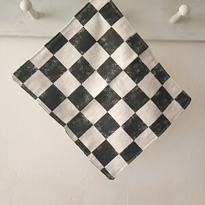 May include: A square, black and white checkered cloth, possibly a kitchen towel or napkin, is displayed against a white background. The fabric has a textured appearance, with the black squares slightly distressed. The edges are neatly sewn.