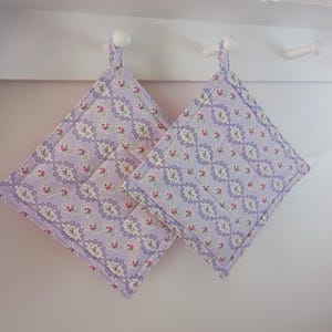 May include: Two square lavender pot holders with a floral pattern of pink and white flowers. The pot holders are hanging on white hooks. The edges are trimmed with a lavender border.