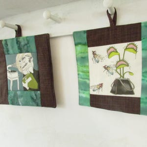 May include: Two square fabric wall hangings with brown and green borders. One features a person listening to a record player, the other a Venus flytrap with flies. Both have brown hanging loops.