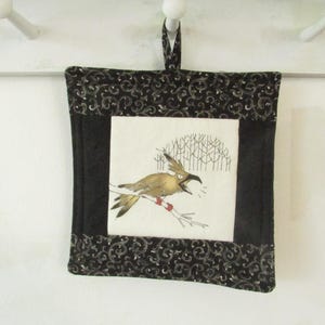 May include: Square, quilted wall hanging featuring a bird illustration. The bird is perched on a branch with a red and white stripe. The artwork is framed by a black border with a swirling pattern and a black backing.