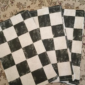 May include: A stack of three square, black and white checkered placemats. The placemats have a distressed, textured appearance and are edged with a white zig-zag stitch. The background is a floral pattern.