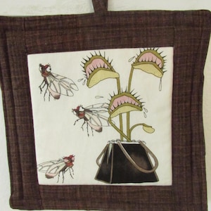 May include: Brown square potholder with a stitched border and a loop for hanging. The center features a whimsical illustration of a Venus flytrap plant with flies. The flytrap has pink and green details, and the flies are red and white.