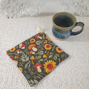 May include: A square quilted potholder featuring a dark green background with a pattern of orange mushrooms, yellow sunflowers, and green leaves, accompanied by a ceramic mug with a blue and green gradient glaze and a sturdy handle.